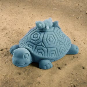 Puede incluir: Una escultura de tortuga de cerámica azul con una tortuga azul más pequeña en su espalda. La tortuga más grande tiene un caparazón detallado con un patrón hexagonal.