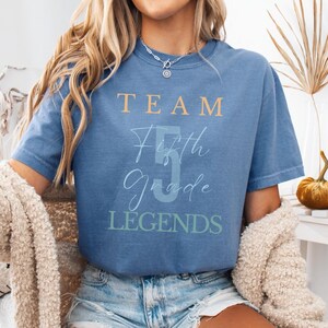 Puede incluir: Camiseta azul vaquero con el texto "TEAM Fifth Grade LEGENDS" en una mezcla de fuentes y colores. La camiseta es de manga corta y tiene un corte relajado. La imagen también incluye una calabaza decorativa y una cesta tejida.