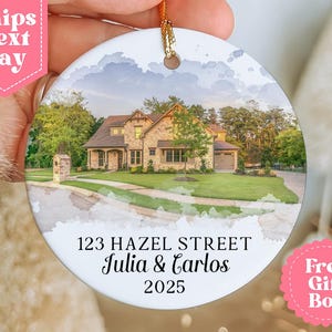 May include: A round ceramic ornament featuring a watercolor image of a house with a green lawn and trees. The ornament has the text "123 HAZEL STREET Julia & Carlos 2025". It has a gold string for hanging.
