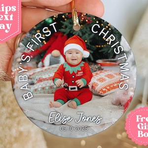 May include: Round Christmas ornament featuring a baby in a red elf outfit and Santa hat. The ornament reads "BABY'S FIRST CHRISTMAS" and includes the name "Elize Jones" and the date "09 10 2025". The ornament has a gold string for hanging.