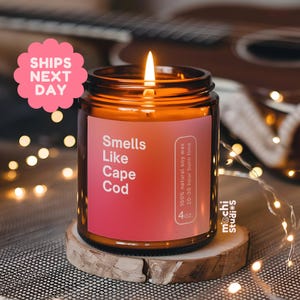 May include: A lit candle in a brown glass jar with a label that reads "Smells Like Cape Cod." The candle sits on a wooden coaster. The label also states the candle is 4 oz and made with 100% natural soy wax.
