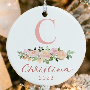 May include: A white ceramic Christmas ornament with a pink floral design and the letter "C" in pink. The ornament also has the name "Christina" and the year "2023" in pink.