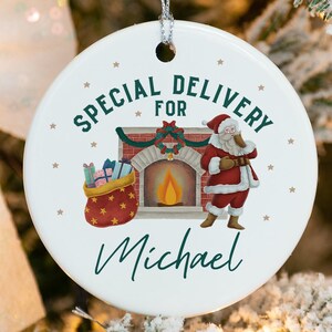 May include: White ceramic Christmas ornament with a red and white Santa Claus illustration, a fireplace, and the text "Special Delivery for Michael".