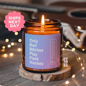 May include: A lit soy wax candle in a glass jar with a label that reads "Only Bad Bitches Play Field Hockey." The candle sits on a wooden coaster. The candle is 4 oz and has a 20-30 hour burn time.