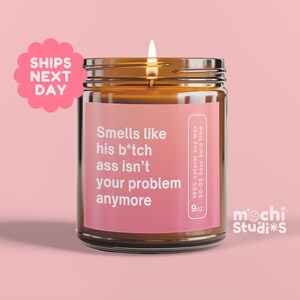 May include: A lit candle in a glass jar with a pink label that reads "Smells like his b*tch ass isn't your problem anymore." The candle is 9 oz and made of 100% natural soy wax with a 50-60 hour burn time. The background is pink.