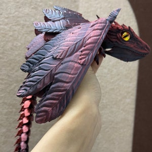 Puede incluir: Una figura de drag&oacute;n impresa en 3D con alas rojas y negras, una cola puntiaguda y un ojo amarillo. El drag&oacute;n est&aacute; en posici&oacute;n de vuelo.