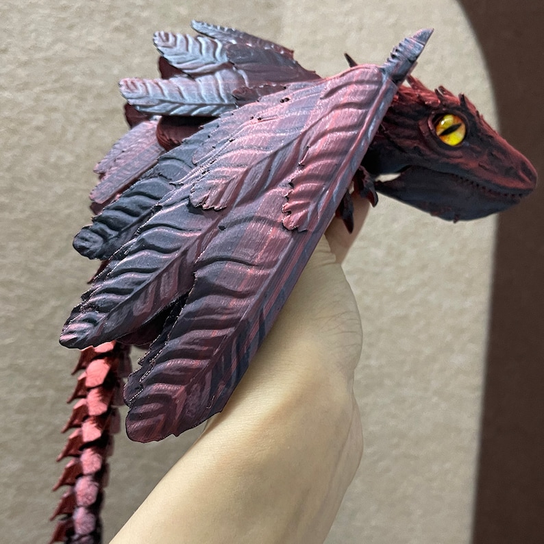 Puede incluir: Una figura de drag&oacute;n impresa en 3D con alas rojas y negras, una cola puntiaguda y un ojo amarillo. El drag&oacute;n est&aacute; en posici&oacute;n de vuelo.
