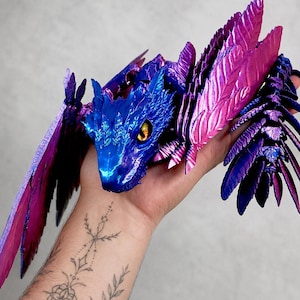 May include: A 3D printed articulated dragon figure featuring a metallic finish with a color-shifting gradient of deep blue and vibrant magenta. The creature has detailed scale textures, large feathered wings, and a single visible yellow eye.