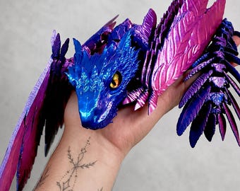 May include: A 3D printed articulated dragon figure featuring a metallic finish with a color-shifting gradient of deep blue and vibrant magenta. The creature has detailed scale textures, large feathered wings, and a single visible yellow eye.