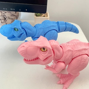 May include: Two toy Tyrannosaurus Rex dinosaurs, one light blue and one pink, are displayed. The dinosaurs have articulated limbs and open mouths, with green and yellow eyes. The toys are on a white surface.