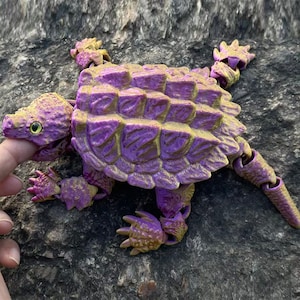 May include: A purple and gold articulated toy ankylosaurus dinosaur. The toy features a textured, segmented body, a small head with a green eye, and articulated limbs. The dinosaur is resting on a rock surface.