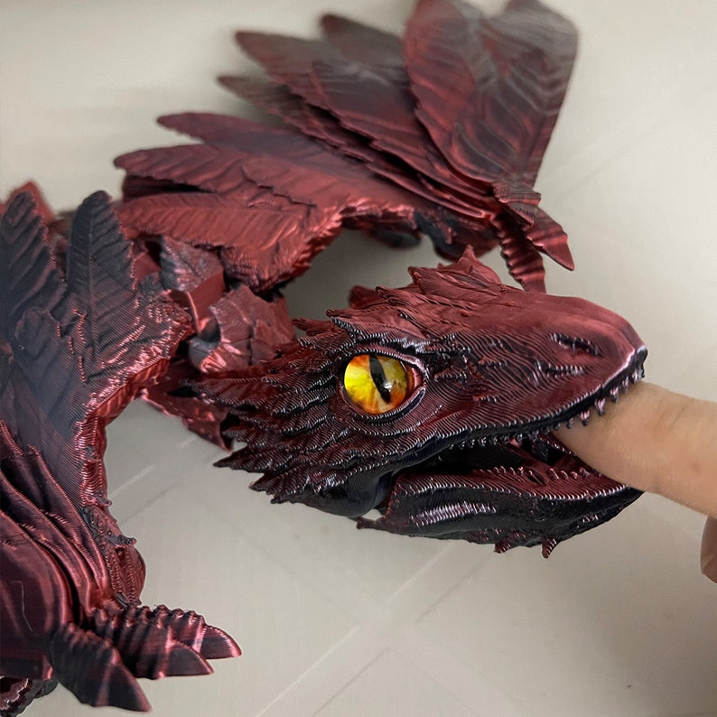 Puede incluir: Una figura de drag&oacute;n roja y negra con un ojo amarillo, la boca abierta y un dedo dentro. El drag&oacute;n tiene escamas y alas detalladas.