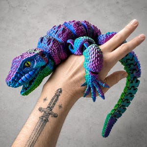 May include: An articulated lizard figurine featuring a vibrant gradient of purple, blue, and green colours. The creature has a textured, scaly surface, a detailed head with visible eyes, and a long, segmented tail that curves downwards.