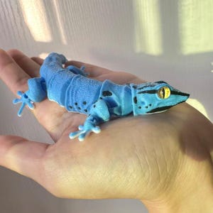 May include: A light blue articulated gecko toy with black spots and stripes. The gecko has a detailed head with yellow eyes and is resting on a hand. The toy is made of plastic and has a segmented body.