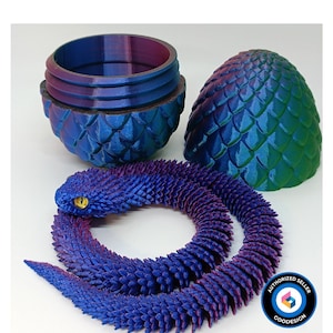 May include: A 3D-printed articulated snake in blue and purple, with a yellow eye, coiled near a matching egg-shaped container and a separate eggshell. The container is in two parts, with a textured, scaled design.