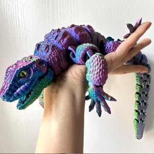 Peut inclure: Un jouet lézard articulé multicolore dans les tons de violet, de bleu et de vert. Le jouet a une surface texturée et détaillée et est tenu dans une main. Les yeux du lézard sont d'un vert-jaune vif.