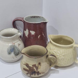 May include: Four ceramic jugs of different sizes and designs. One is burgundy with white accents, another is cream-coloured with a floral pattern, and a third is beige with brown brushstrokes. The fourth jug is white with a blue and grey design.