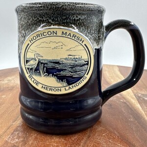 May include: A ceramic mug with a dark blue base and a mottled grey rim. A circular emblem on the front features a black and white illustration of a heron, a boat, and the text Horicon Marsh Blue Heron Landing in a circular border.
