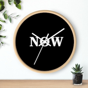 May include: A black and white clock with a wooden frame. The clock face has the word "NOW" printed in white on a black background.