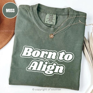 May include: A moss green t-shirt with the words "Born to Align" in white, bold, retro-style lettering. The shirt has a crew neck and short sleeves. A gold necklace with a pendant is draped across the shirt.