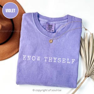 Può includere: Una maglietta Comfort Colors color lavanda con la scritta "KNOW THYSELF" in bianco. Una collana d'oro con un ciondolo è appoggiata sulla maglietta. Un cappello marrone e una pianta secca sono sullo sfondo.