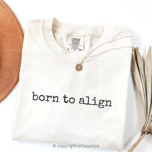 Puede incluir: Camiseta color crema con la frase "born to align" impresa en negro. Un collar dorado con un colgante circular descansa sobre la camiseta. La camiseta es de la marca Comfort Colors, talla S. Un sombrero marrón y una planta seca en el fondo.