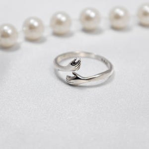 May include: A silver ring with two hands clasped together, forming a circle. The ring is on a white background.