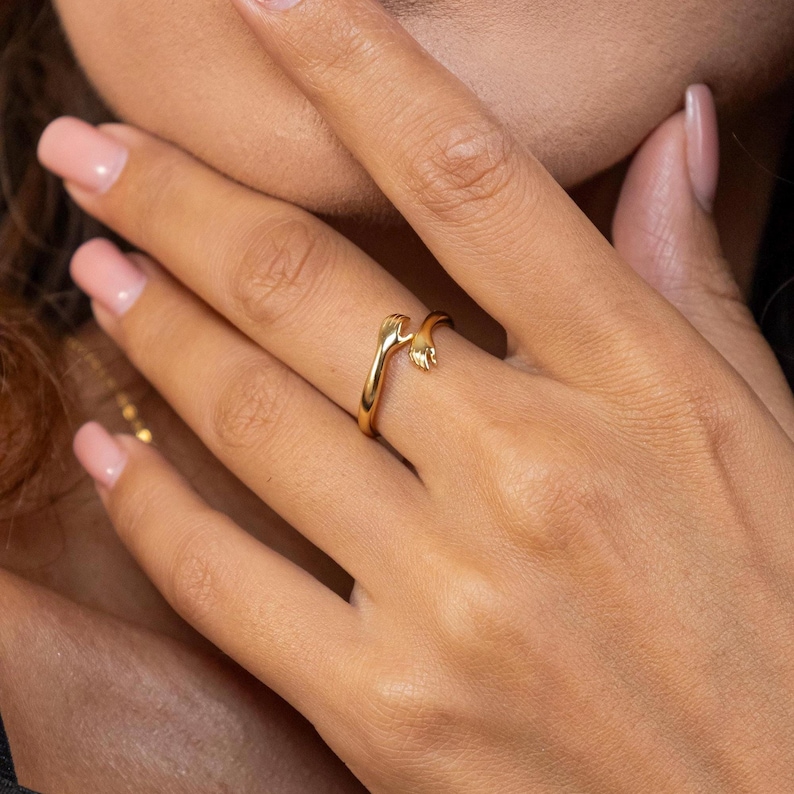 May include: A gold ring with two hands clasped together, the ring is worn on a person's finger.