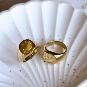 May include: Two gold signet rings featuring a smiley face design. The rings are presented on a white, shell-shaped dish. The rings have a polished finish and a rounded shape. The smiley face is embossed on the face of each ring.