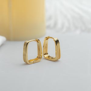May include: A pair of gold-coloured, square hoop earrings. Each earring has a layered design with three parallel bars forming the hoop. The earrings are displayed on a light-coloured surface, with a soft, blurred background.