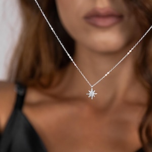 May include: A silver necklace with a starburst pendant. The pendant is made of small, sparkling stones. The necklace is delicate and hangs on a thin chain. The background is blurred, focusing on the necklace.