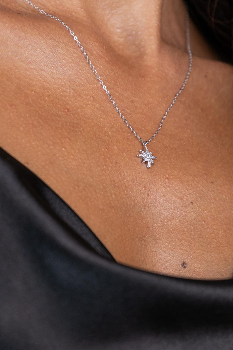 May include: A silver necklace with a starburst pendant. The pendant is encrusted with small, sparkling stones. The necklace is displayed on a person's neck, against a black background.