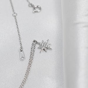 May include: A delicate silver necklace with a starburst pendant. The pendant is encrusted with small, sparkling stones. The necklace is displayed on a white, textured background, highlighting the jewelry's shine and intricate design. The chain has a clasp and an adjustable length.