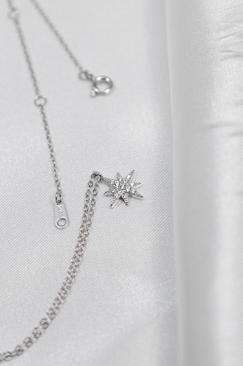 May include: A delicate silver necklace with a starburst pendant. The pendant is encrusted with small, sparkling stones. The necklace is displayed on a white, textured background, highlighting the jewelry's shine and intricate design. The chain has a clasp and an adjustable length.