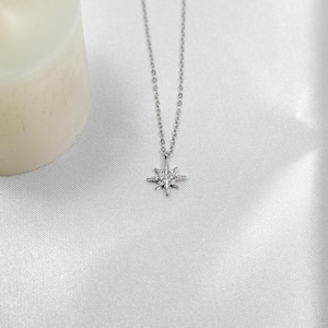 May include: A silver-colored necklace with a starburst pendant. The pendant is detailed with small, sparkling accents. The necklace hangs on a delicate chain, and the background is a soft, white fabric.