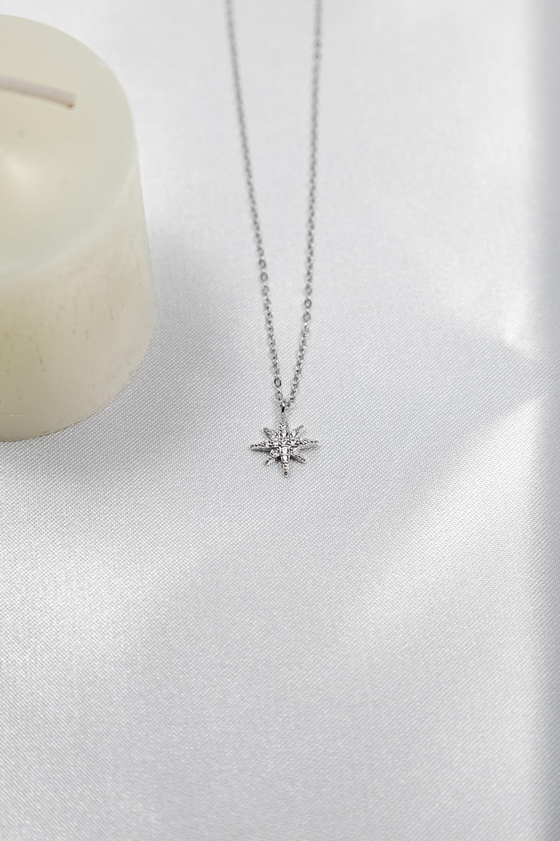 May include: A silver-colored necklace with a starburst pendant. The pendant is detailed with small, sparkling accents. The necklace hangs on a delicate chain, and the background is a soft, white fabric.