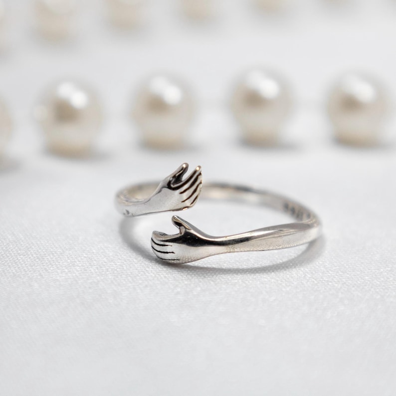 May include: A silver ring with two hands clasped together, forming a circle. The ring is designed to look like two hands embracing each other.