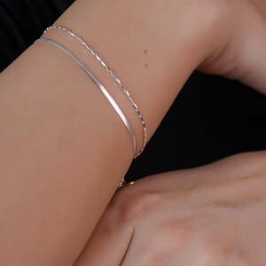 May include: Two silver bracelets on a person's arm. The bracelets are both thin and delicate, one is a simple band and the other is a chain.