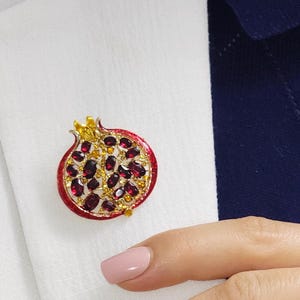 May include: A pomegranate brooch with a red enamel border, gold accents, and ruby-colored stones. The brooch is attached to a white shirt. The brooch is a jewelry item.