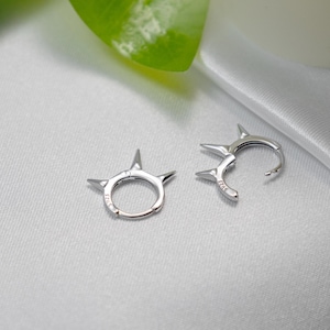 May include: A pair of silver hoop earrings with a pointed, spiky design. The earrings are made of metal and have a shiny finish.