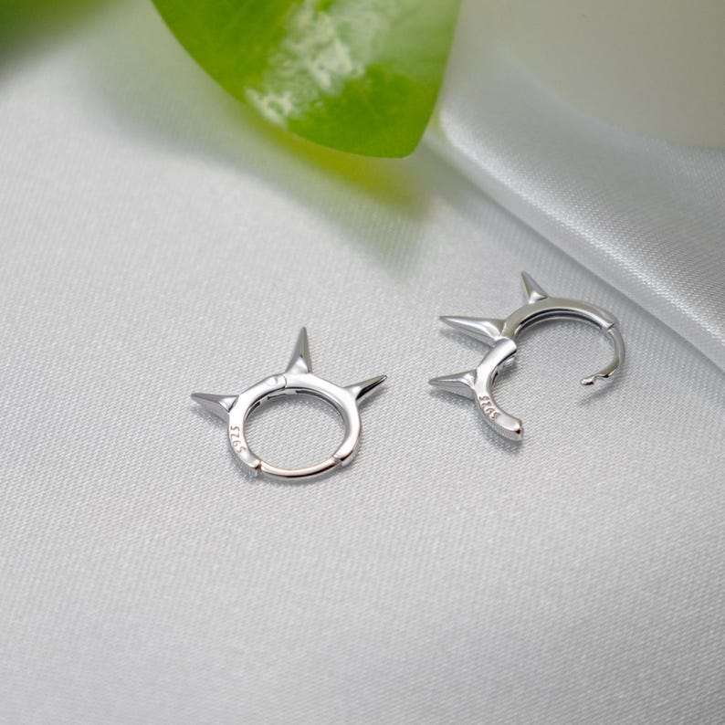 May include: A pair of silver hoop earrings with a pointed, spiky design. The earrings are made of metal and have a shiny finish.
