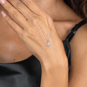 May include: A delicate silver necklace featuring a starburst pendant with small, sparkling accents. The necklace is displayed against a black background, highlighting the pendant's intricate design and the chain's subtle shine. A simple yet elegant piece of jewelry.