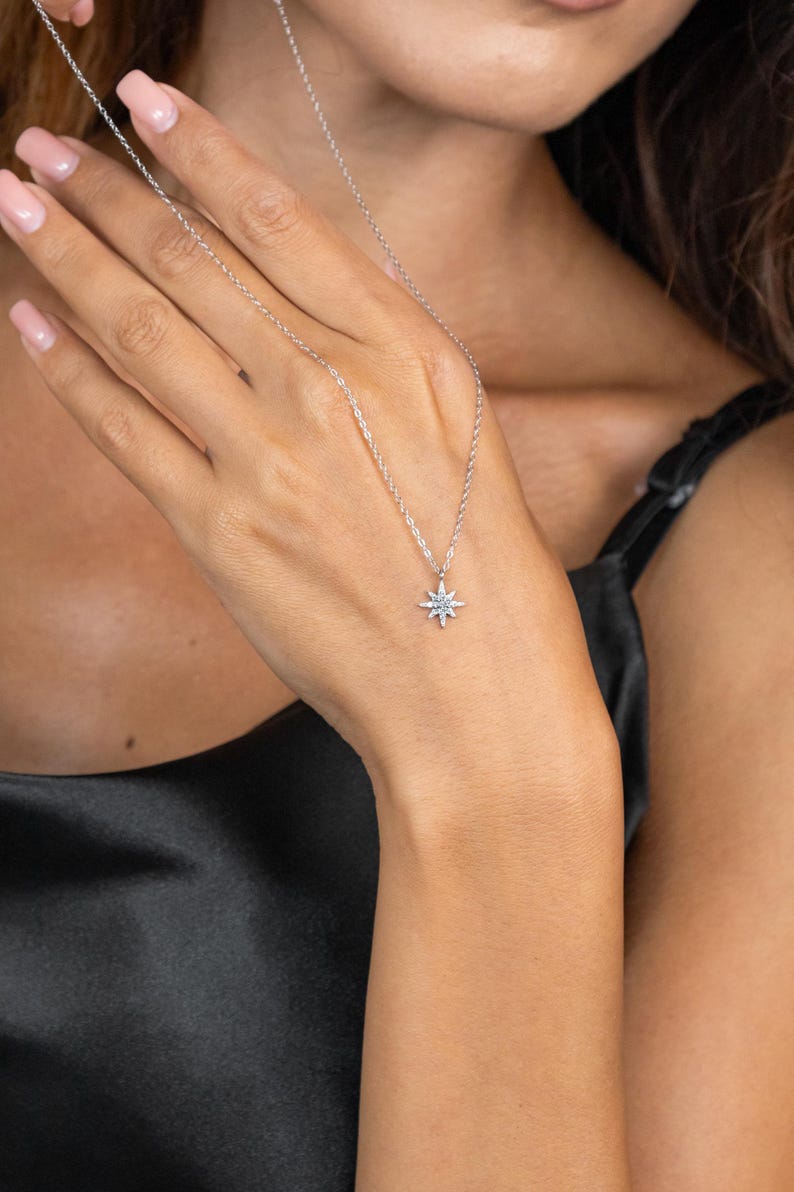 May include: A delicate silver necklace featuring a starburst pendant with small, sparkling accents. The necklace is displayed against a black background, highlighting the pendant's intricate design and the chain's subtle shine. A simple yet elegant piece of jewelry.