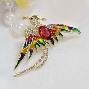 May include: A gold-toned phoenix brooch with a large red gemstone at its center. The wings are adorned with colorful enamel in shades of red, yellow, green, and purple. The body and tail feature clear crystals, adding sparkle to this decorative accessory.
