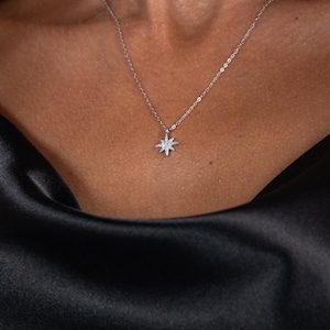May include: A delicate silver necklace featuring a starburst pendant adorned with small, sparkling crystals. The necklace is displayed against a dark, silky fabric, creating a contrast that highlights the pendant's brilliance. The jewelry is a subtle yet elegant accessory.