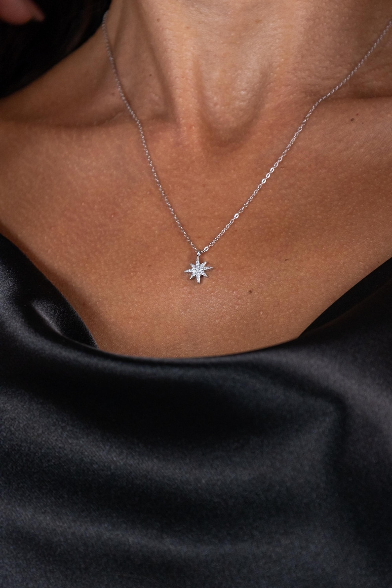 May include: A delicate silver necklace featuring a starburst pendant adorned with small, sparkling crystals. The necklace is displayed against a dark, silky fabric, creating a contrast that highlights the pendant's brilliance. The jewelry is a subtle yet elegant accessory.