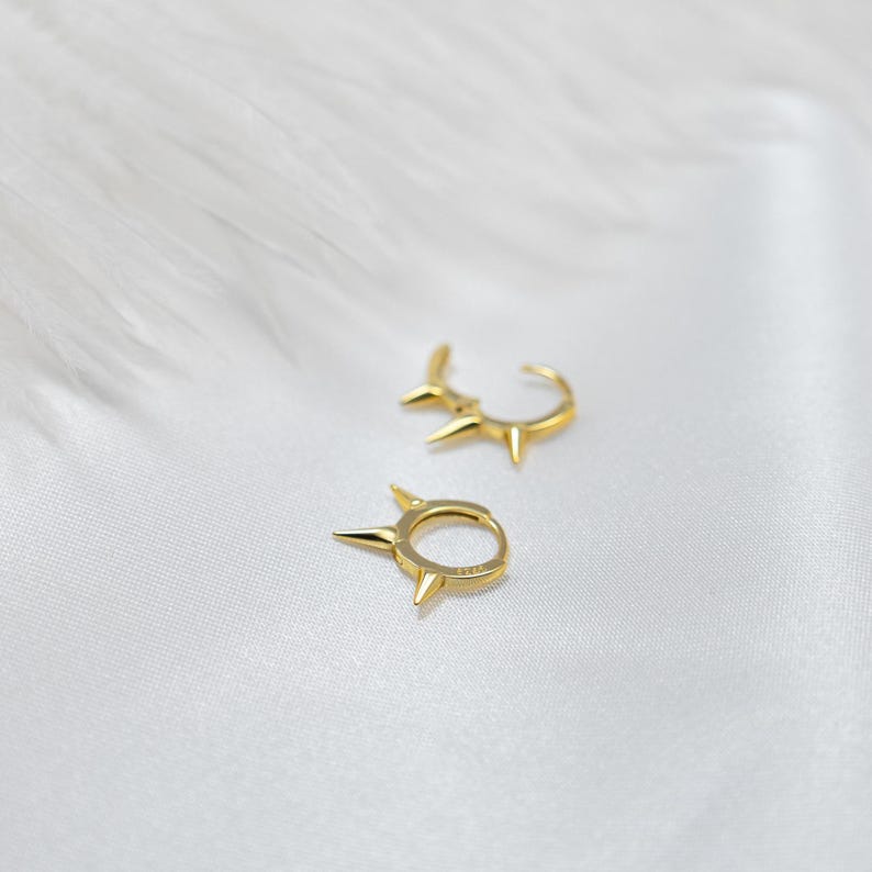 May include: Two gold hoop earrings with a pointed design. The earrings are on a white background.