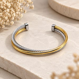 May include: A two-tone cuff bracelet featuring a twisted silver-colored cable strand intertwined with a smooth, polished gold-colored band. The open-ended design terminates in two rectangular silver-colored metal caps at each end of the bracelet.