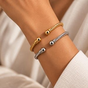 May include: Two open cuff bracelets, one in a polished gold tone and one in a polished silver tone. Each bracelet features a twisted cable texture along the band and is finished with a smooth, spherical ball at each open end of the cuff.