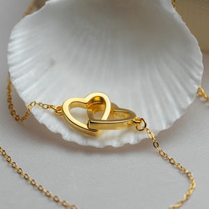 Könnte beinhalten: Goldfarbene Halskette mit zwei ineinandergreifenden Herzanhängern. Die zarte Kette und die Herz-Charms werden auf einer weißen Muschelschale präsentiert, was eine romantische und elegante Ästhetik erzeugt. Ein Symbol für Liebe und Verbundenheit.